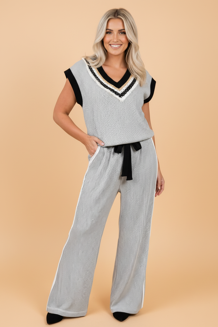 A woman modeling a light gray and black patterned knit lounge set consisting of a sleeveless V-neck top with black trim and matching wide-leg pants with a black tie belt, set against a solid peach background.